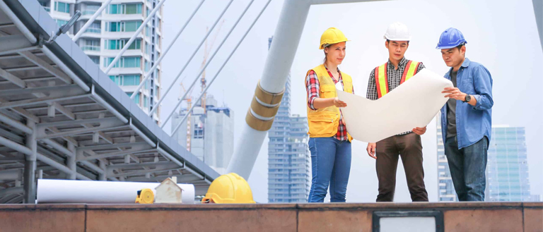 Three students in hard hats standing on a bridge, examining a blueprint for their civil engineering homework and assignment