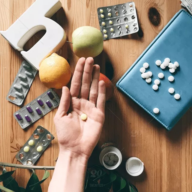 A hand holding a single tablet is surrounded by various pill blister packs, a prescription bottle, fruits, and a blue case on a wooden table—an ideal setting for anyone seeking pharmacology homework help