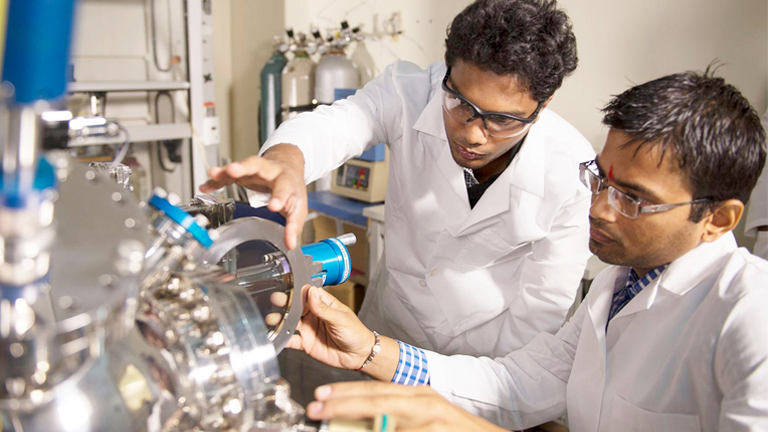 Two chemistry students in lab coats working on a machine, seemingly engrossed in a polymer chemistry assignment